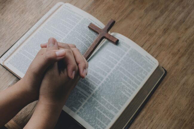 Woman with Bible praying, hands clasped together on her Bible on wooden table.